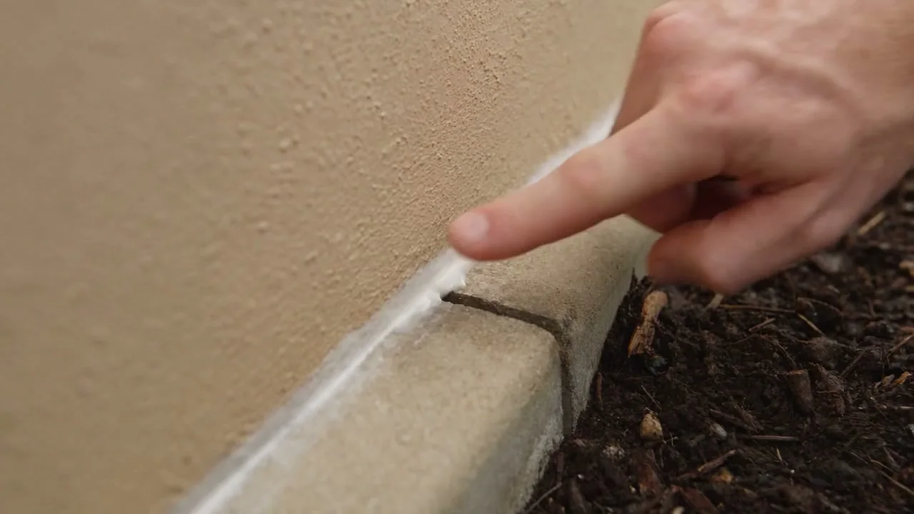 A finger pressed against a freshly run bead of sealant where a rendered wall meets the concrete slab edge
